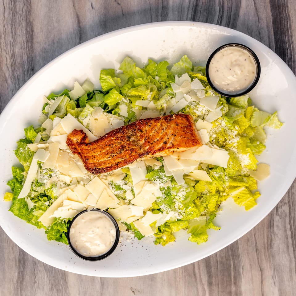 Grilled salmon Caesar salad with parmesan shavings and two cups of dressing on a white platter.
