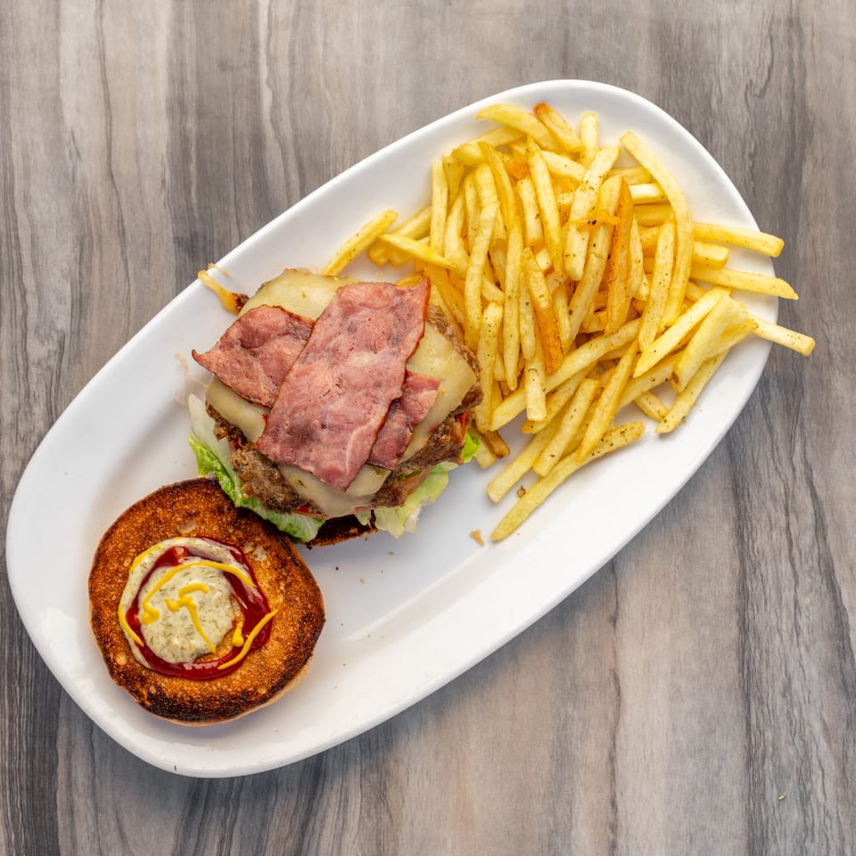 Overhead view of a bacon cheeseburger with fries on a white plate, bun top with condiments.