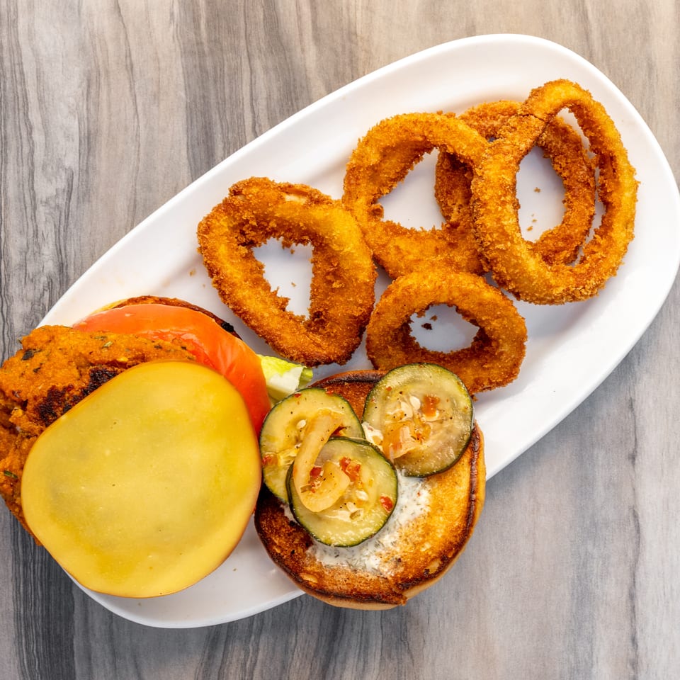 A deconstructed veggie burger with a patty, tomato, cheese, and zucchini on a bun, served with onion rings.